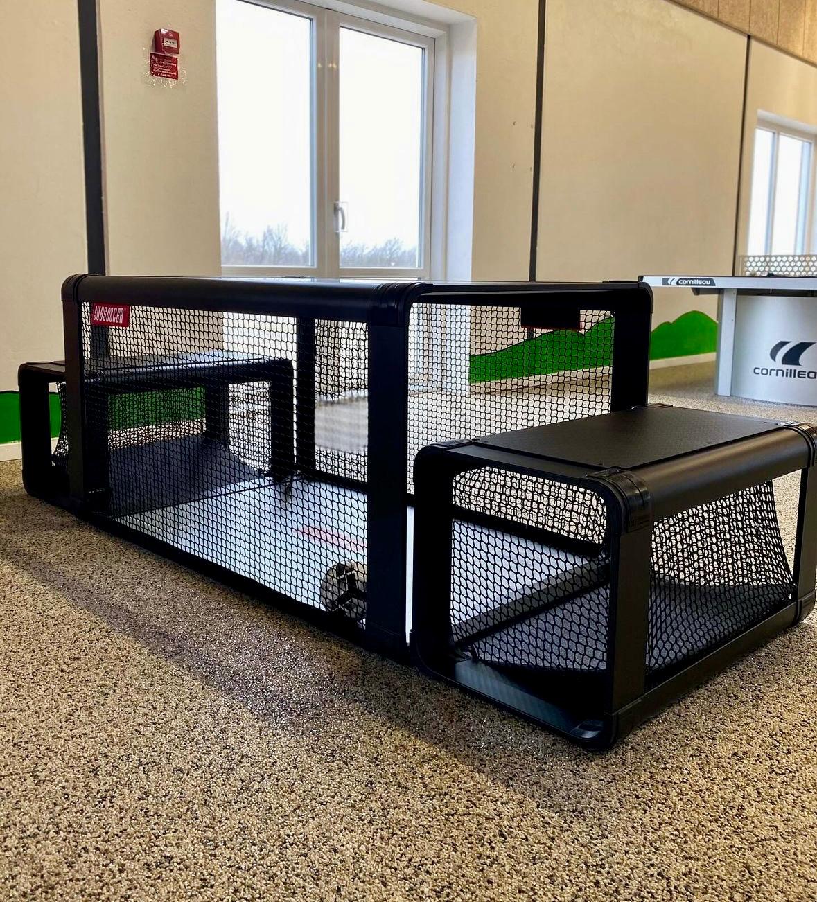 SubSoccer S7 black bench soccer table set up inside a sports hall or leisure centre alongside a ping pong table